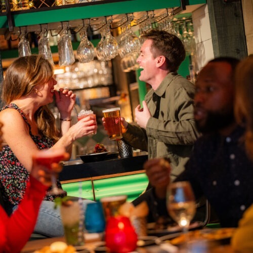 2 Friends Laughing at Bar with Cocktails and Red Stripe 1